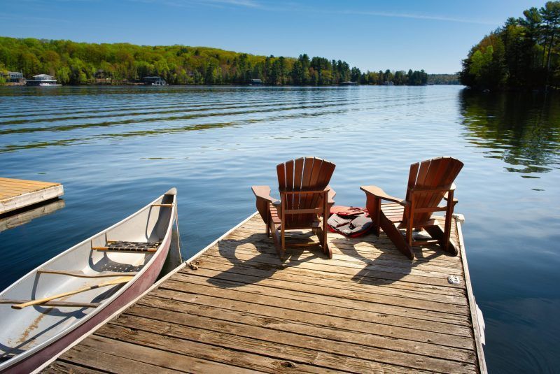 Wooden dock with two Adirondack chairs, a canoe, and a serene lake view with trees and blue sky.
