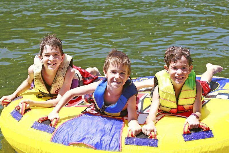 Three children on a yellow inflatable raft, wearing life vests, smiling on a lake.