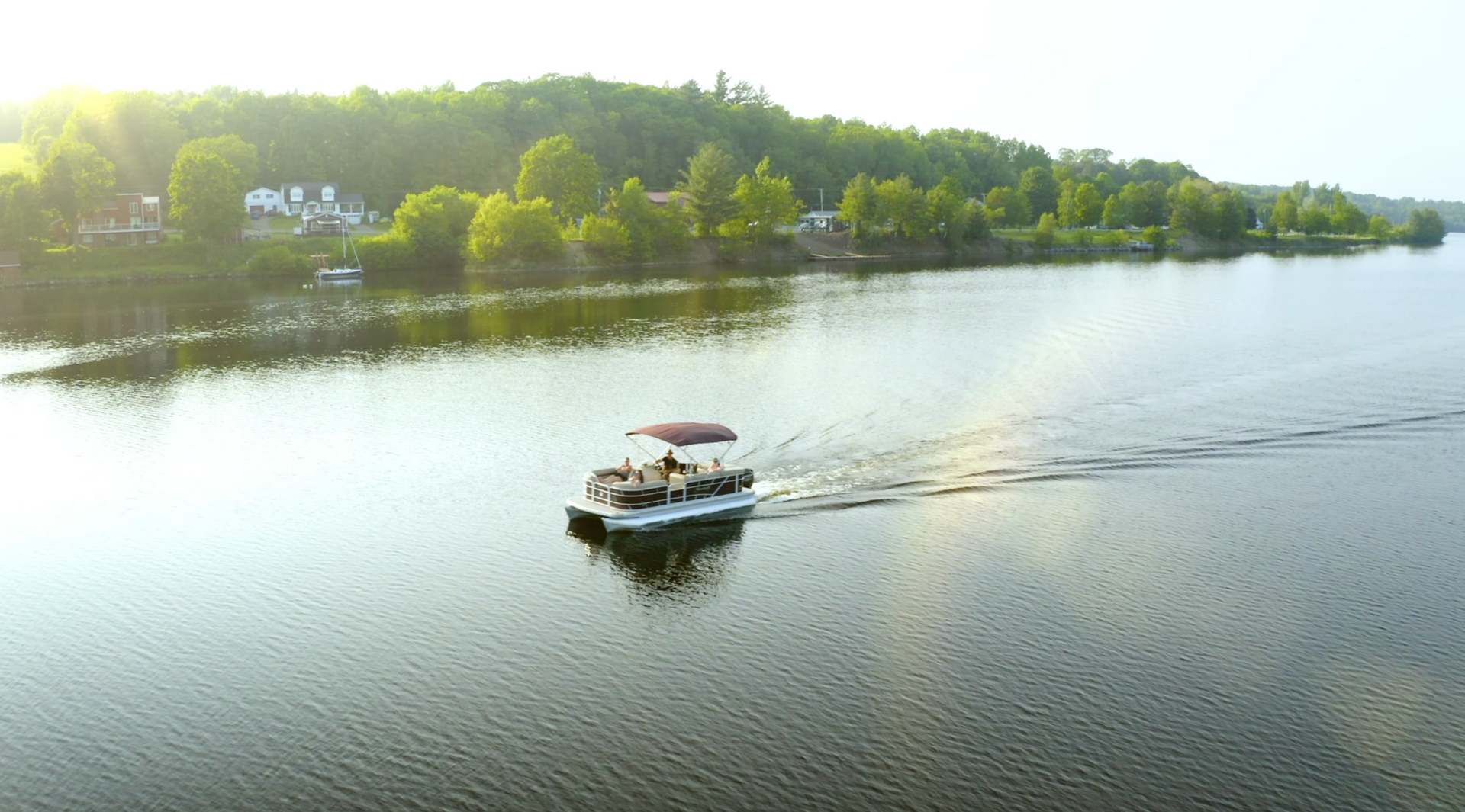 A pontoon boat with a red canopy cruises along a calm, reflective river lined with trees under golden sunlight.