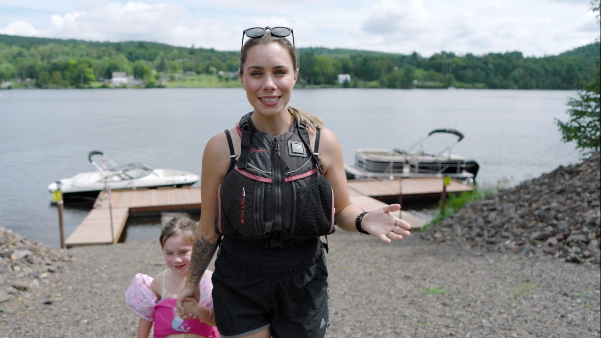 Woman and child by a lake, woman gesturing, wearing life vest, docks and boats in background.