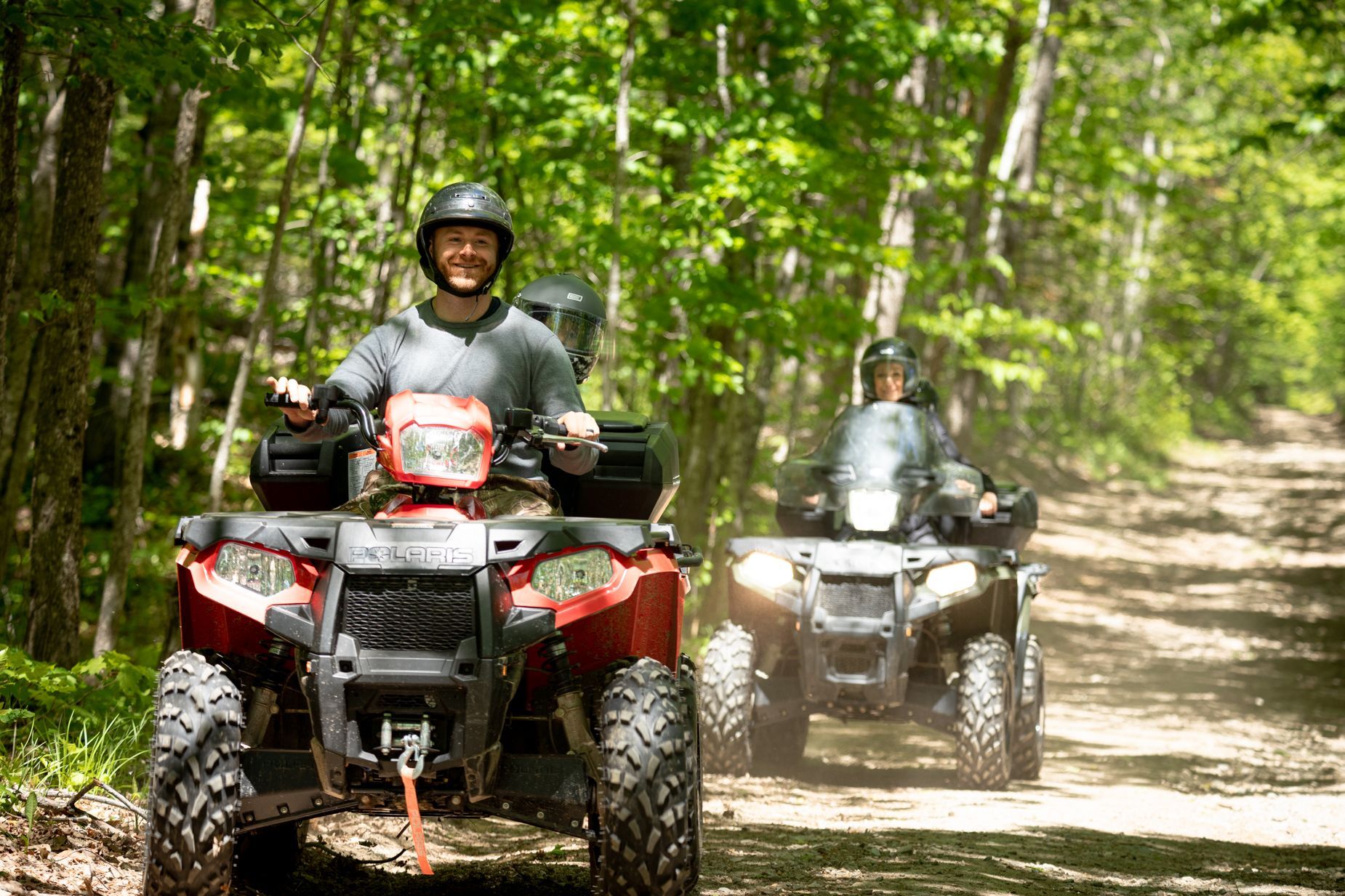 Two people are riding atvs down a dirt road in the woods.