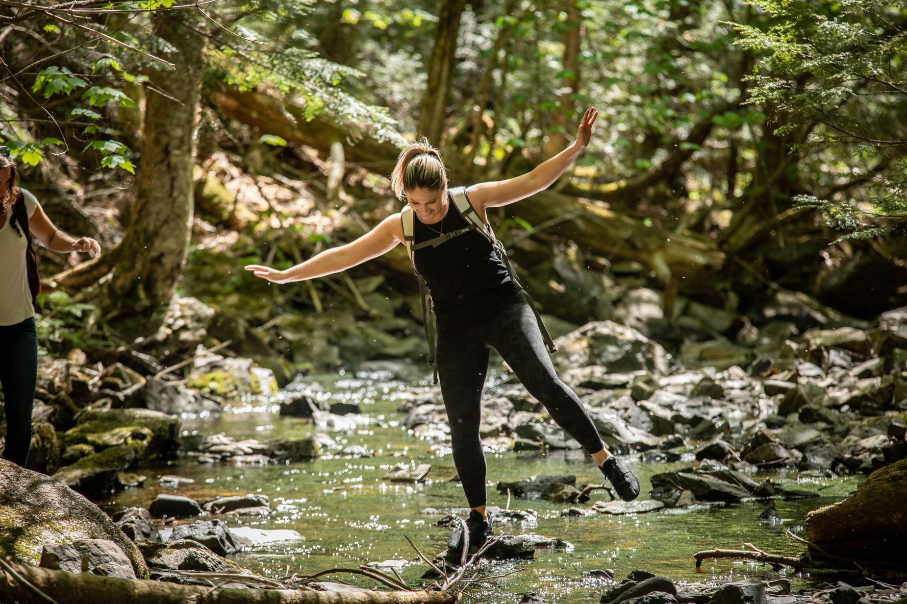 Woman balances on rocks in a stream, arms outstretched for balance. Forest setting.