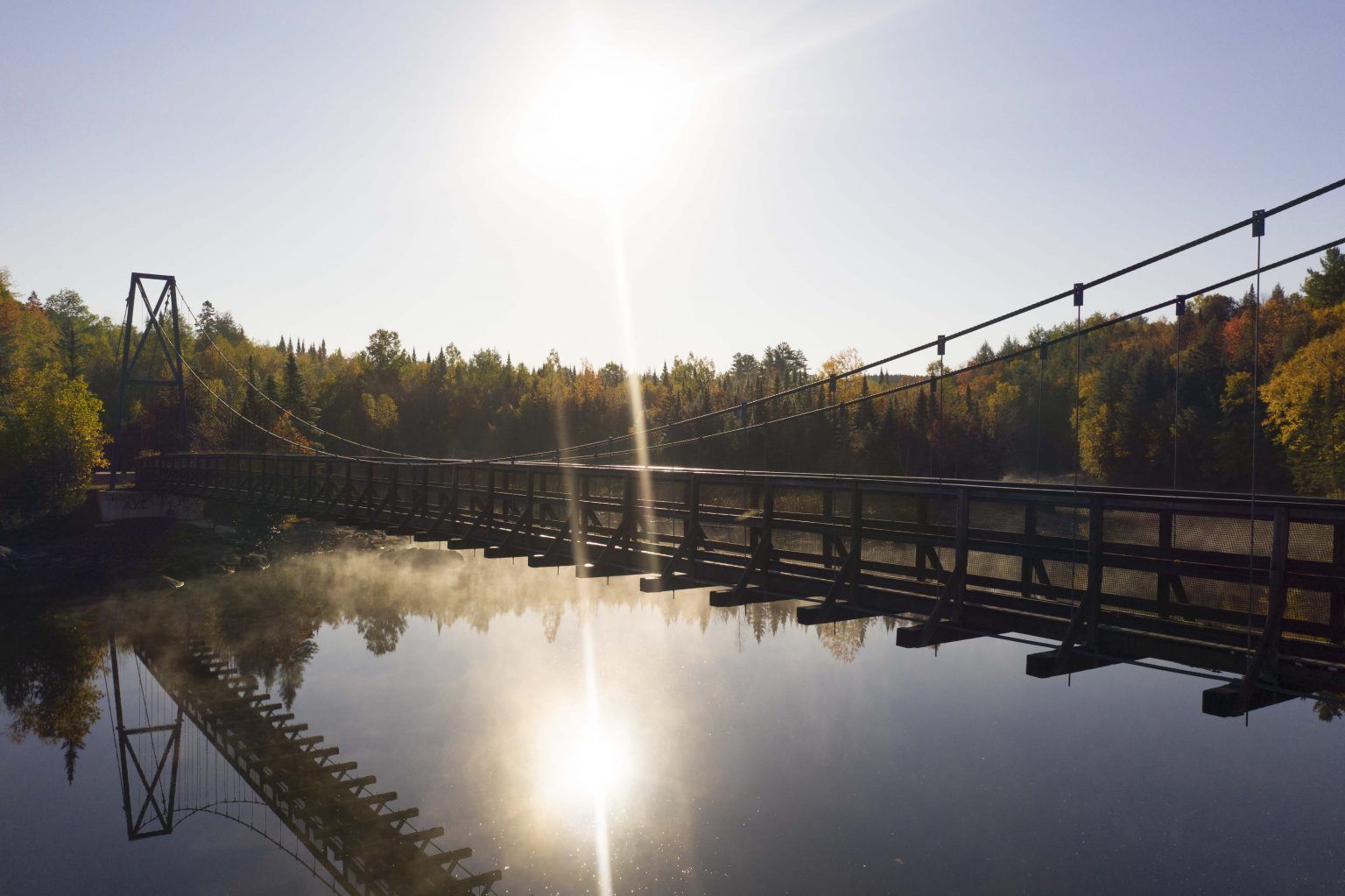 Suspension bridge spanning a river, sun shining brightly. Water reflects the bridge and sky.
