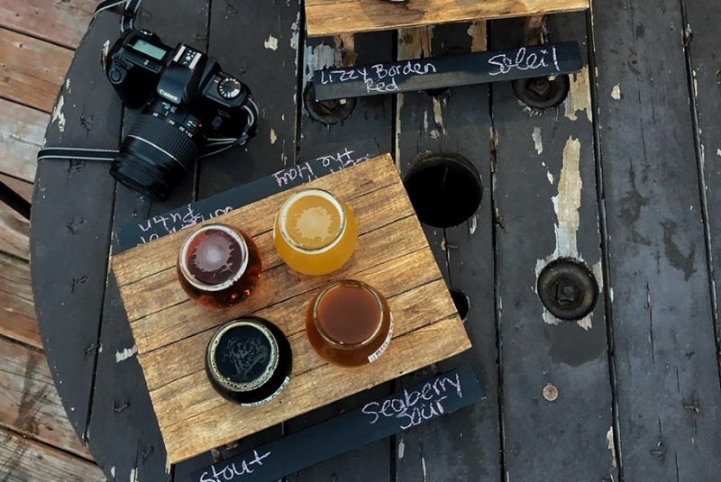 Beer flight with four samples on a wooden plank, next to a camera on a spool table.