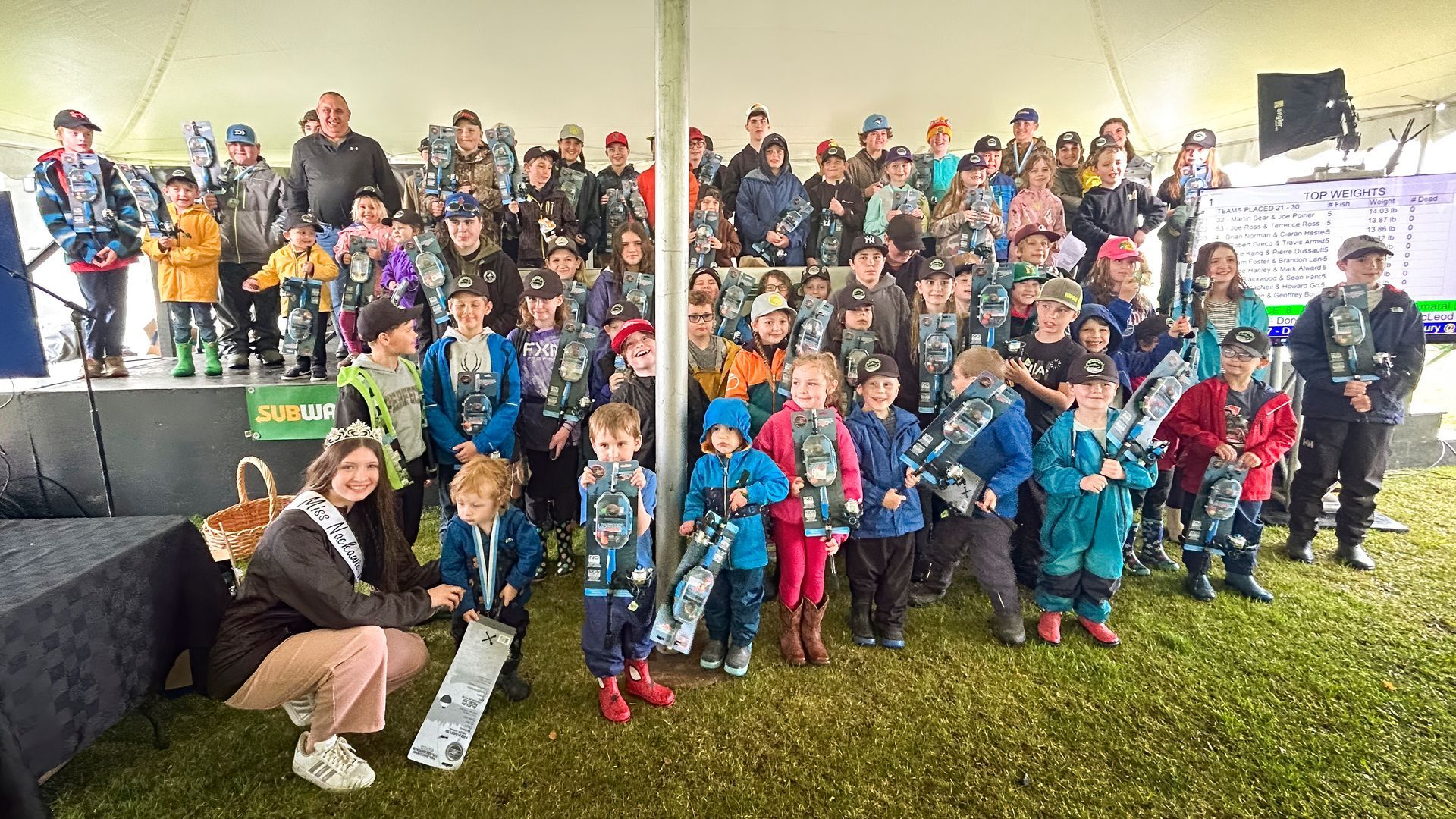 Large group of children and adults posing together outdoors under a tent. Many children wear matching vests.