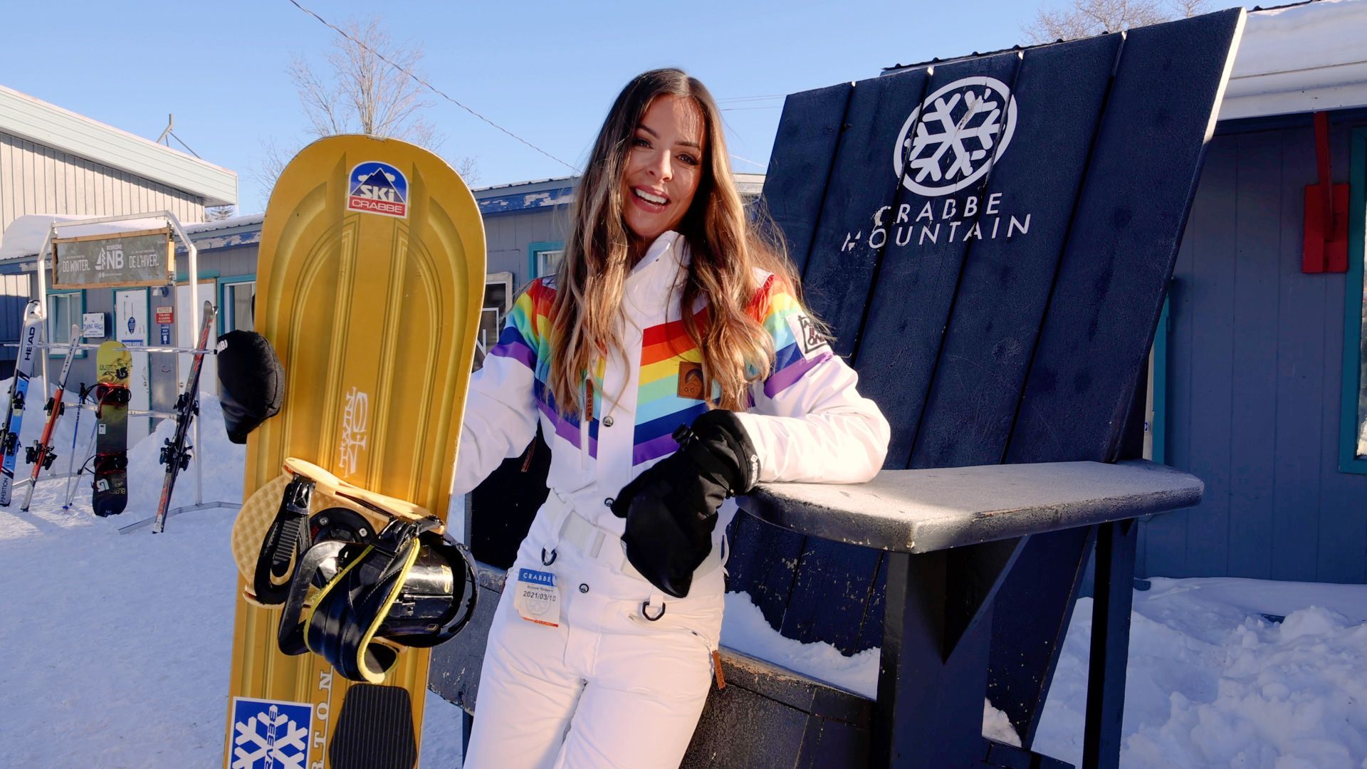 Woman in rainbow jacket stands next to a yellow snowboard and a sign for Frasier Mountain, snowy outdoor setting.