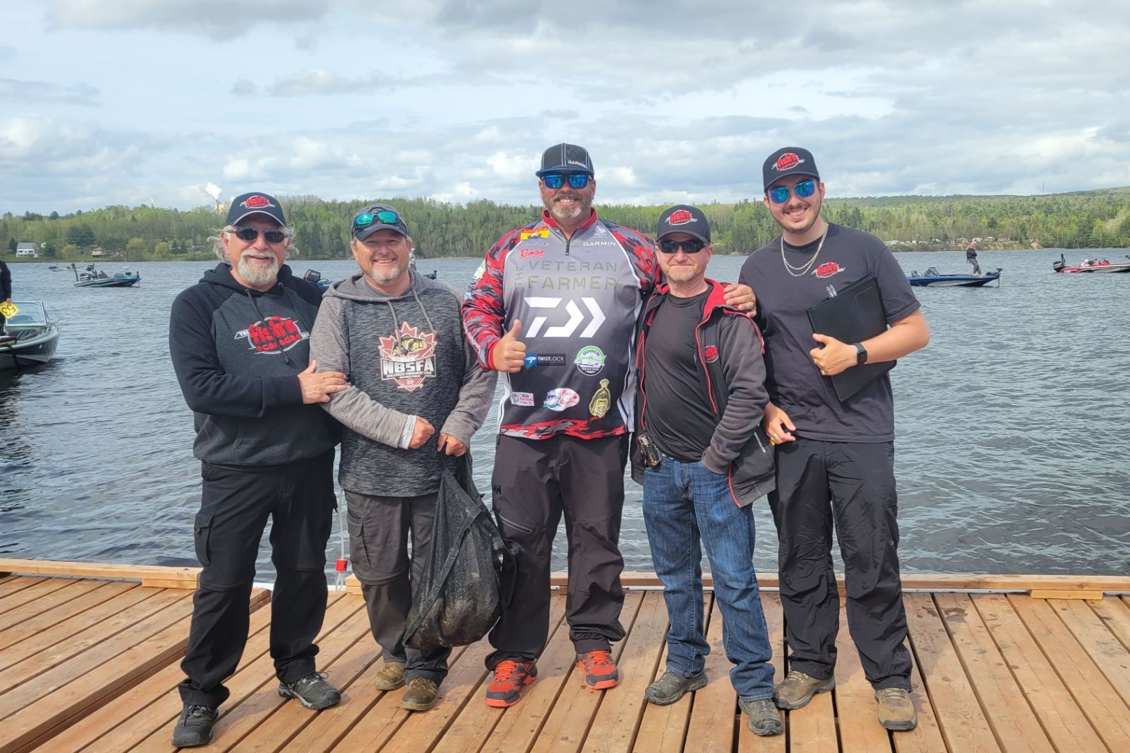 Five people on a dock, lake backdrop. One gives thumbs up, all wear hats, fishing boats in background.