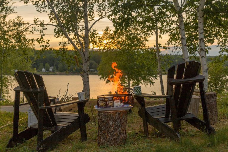 Two Adirondack chairs face a campfire at sunset over a lake.