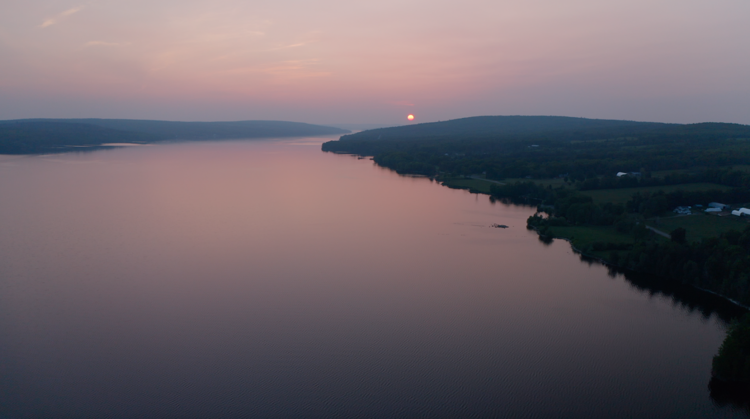 A calm lake reflects the dusky sky, with a sun near the treeline on the distant shore.