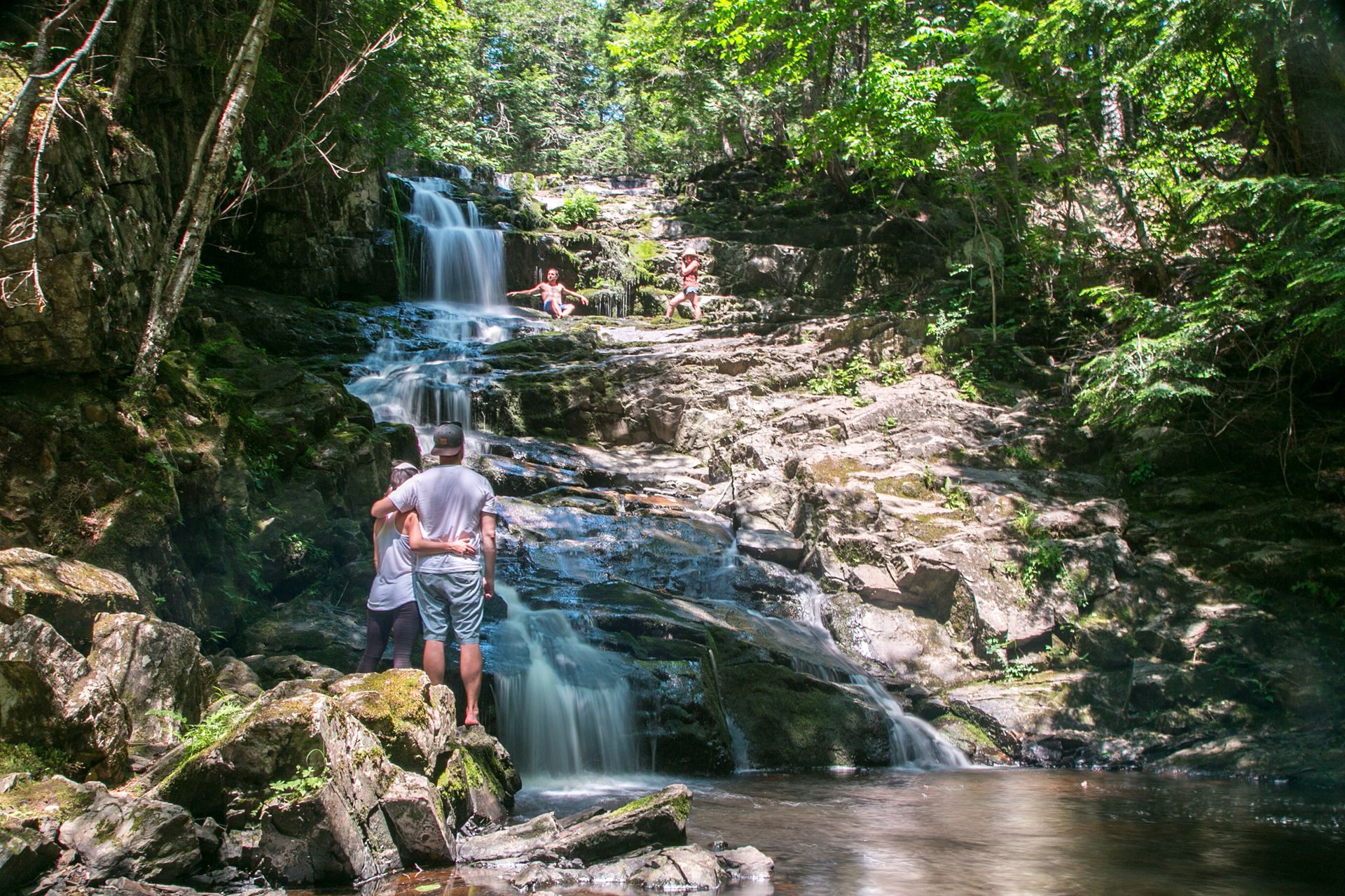 Waterfall cascades over rocks into a pool. People explore the falls in a lush green forest setting.