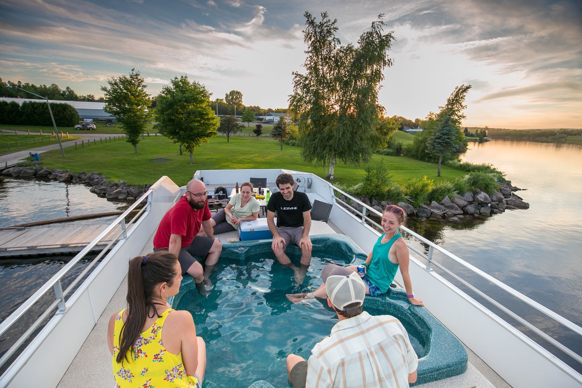 Group of people relaxing in a hot tub on a boat deck at sunset, near a lake.