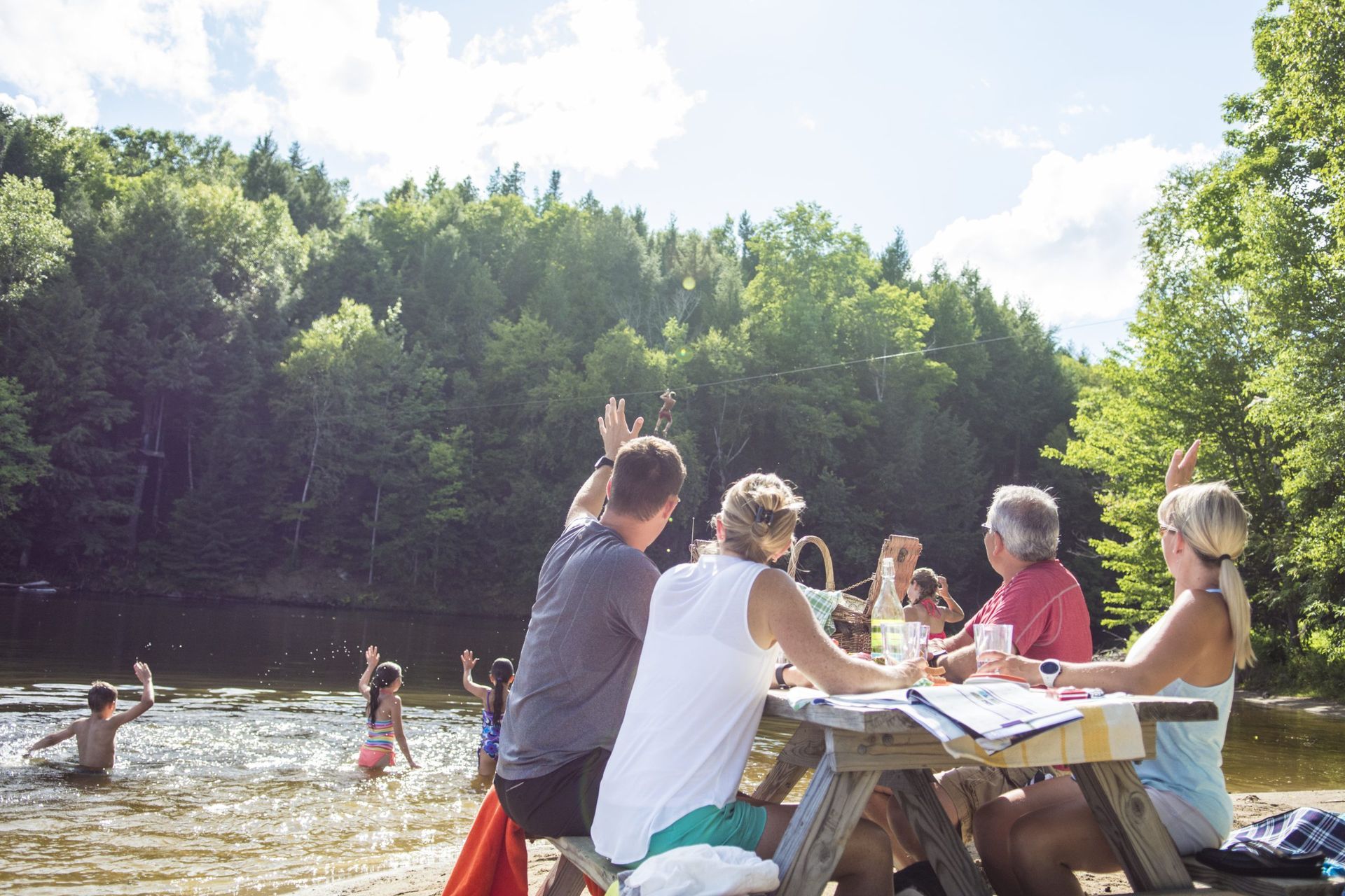 People at a picnic table near a lake, with others playing and splashing in the water. Sunny day.