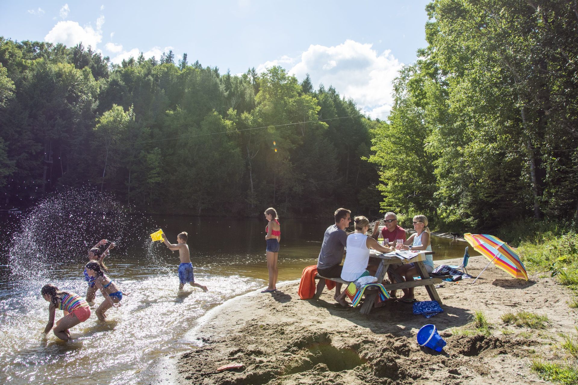 People at a picnic table and playing in the water near a forest.