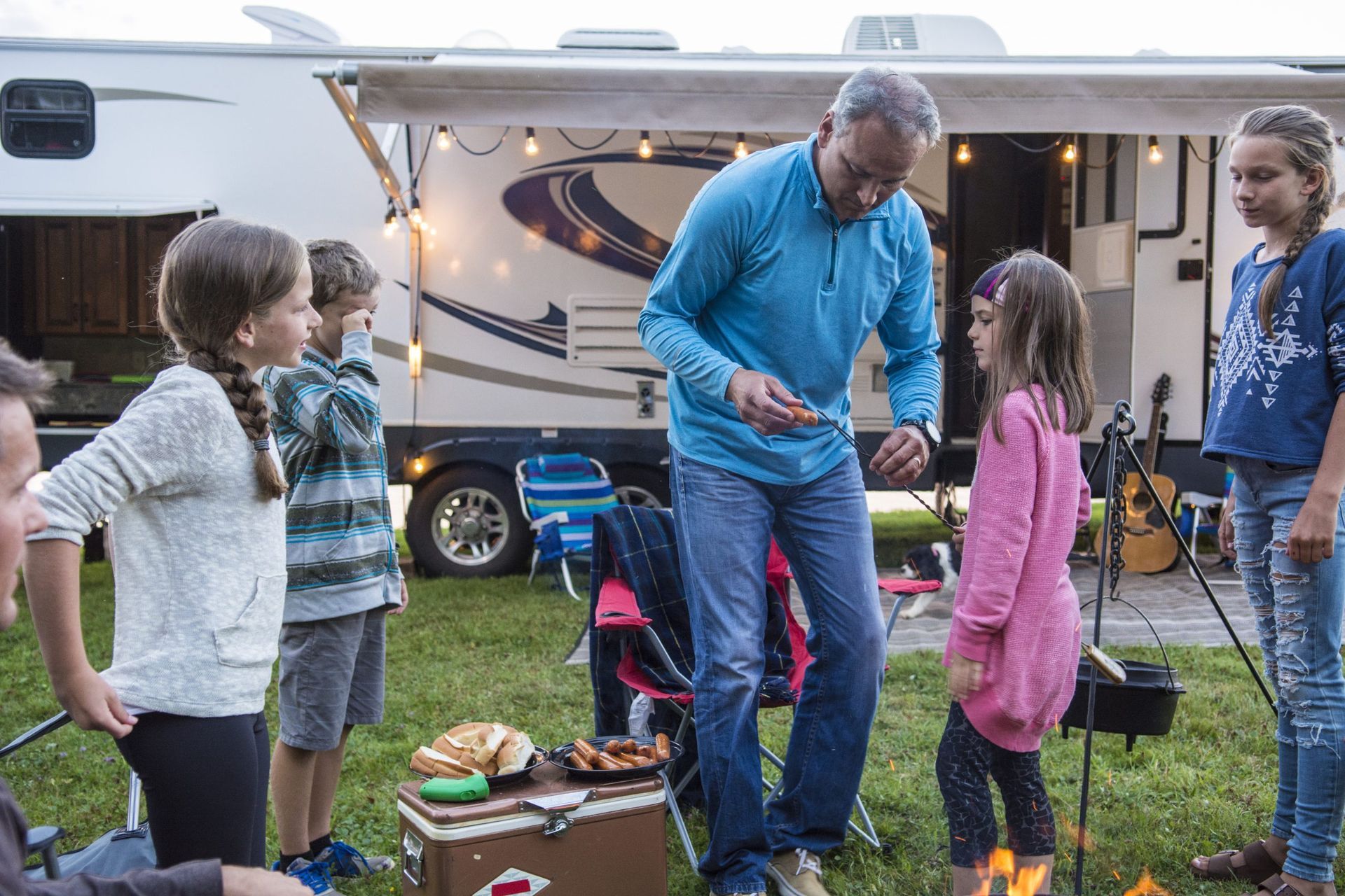 Man shows food to children around a campfire, RV in background.