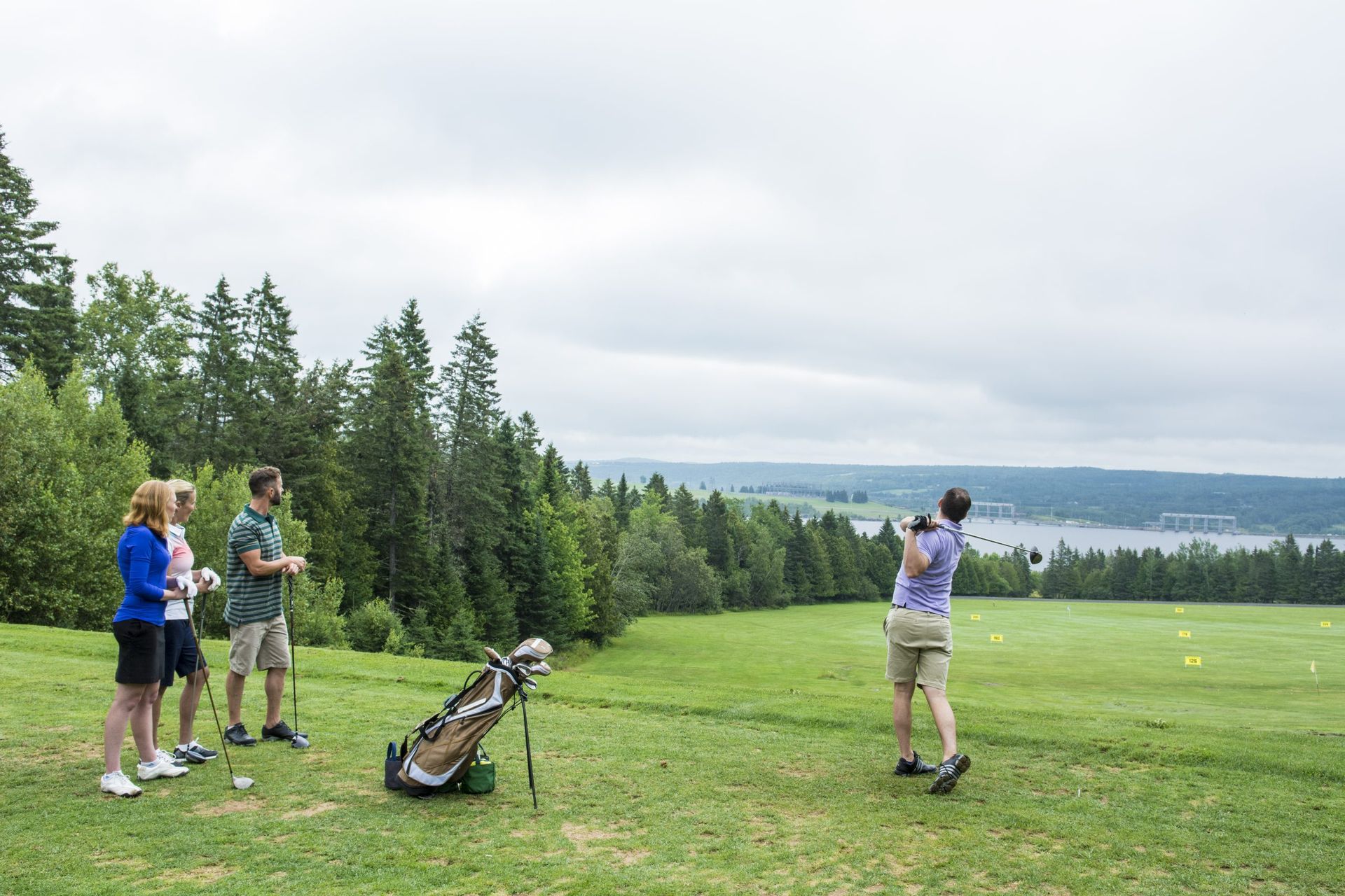 People golfing on a green with lake and trees in the background under a cloudy sky.