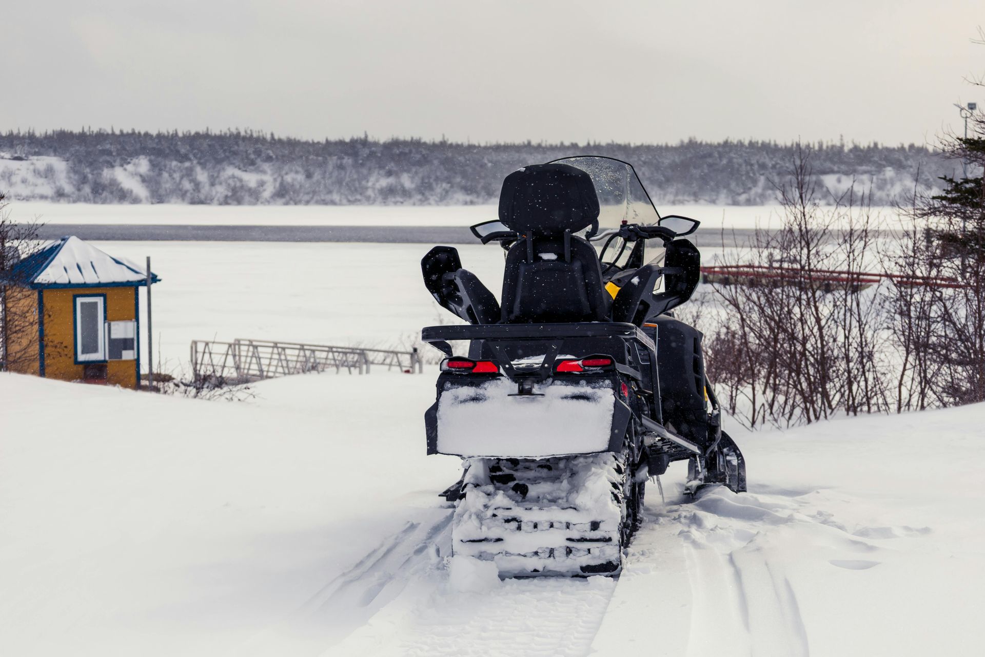 Snowmobile parked on snow, near a body of water. Cabin in the distance. Overcast day.