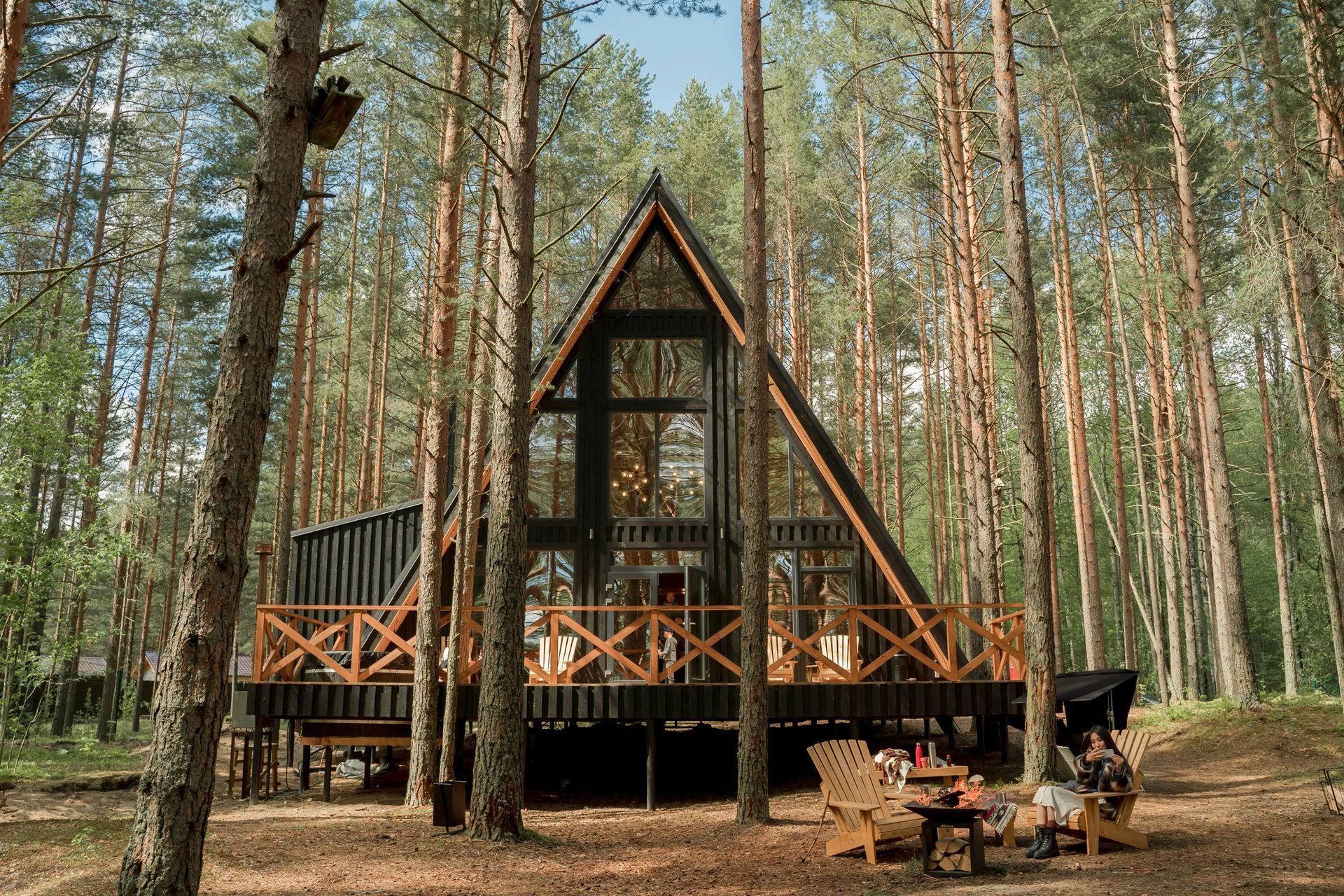 A-frame cabin nestled in a forest with tall trees and a wooden deck, a fire pit, and chairs.