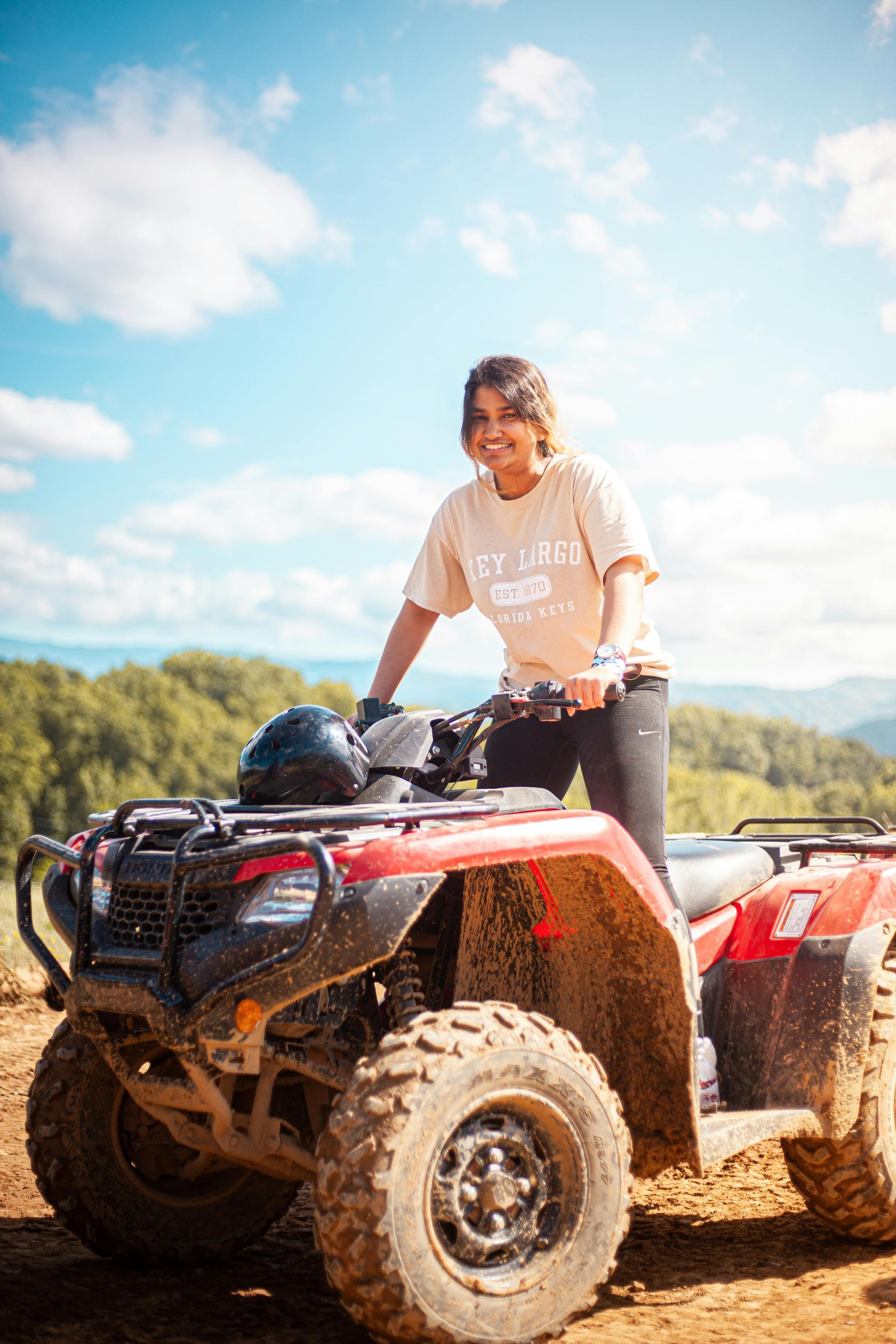Woman smiles while sitting on a dirty red ATV on a mountain with a blue sky in the background.