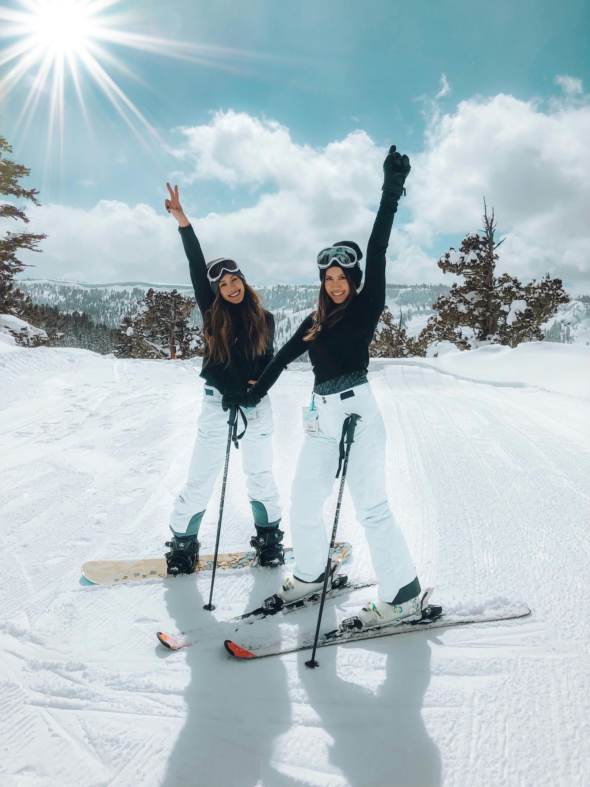 Two people in ski gear on snowy mountain, raising arms in excitement under sunny sky.