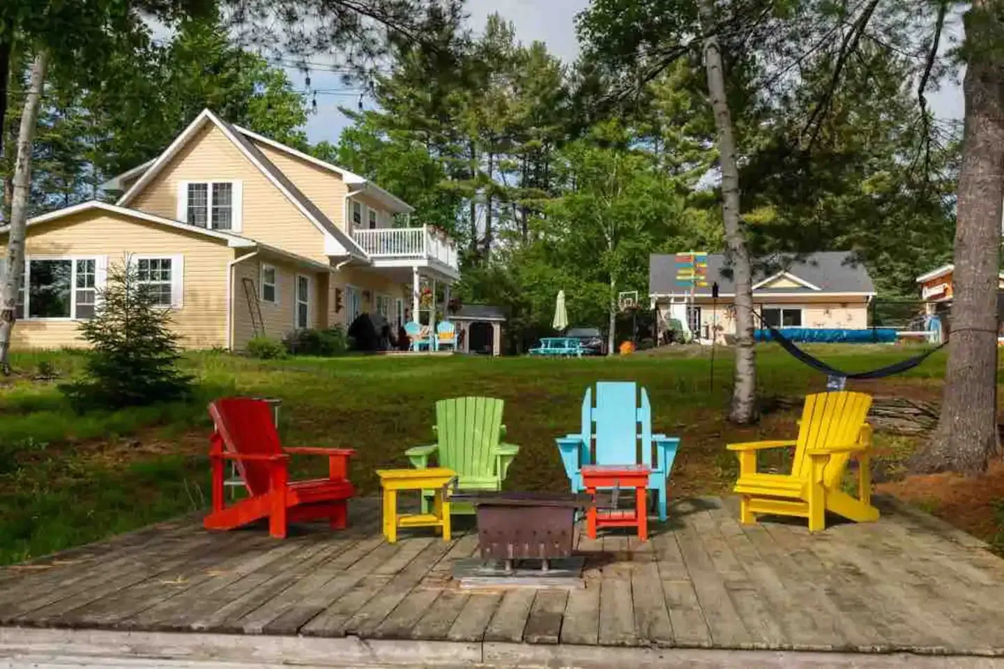 Colorful Adirondack chairs surround a fire pit on a lakeside deck, overlooking a yellow house and trees.