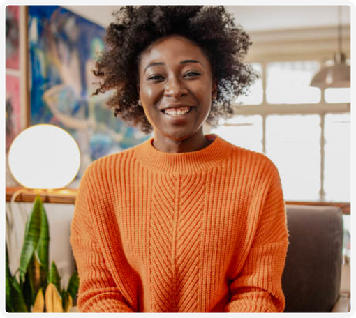 A woman in an orange sweater is smiling while sitting on a couch.