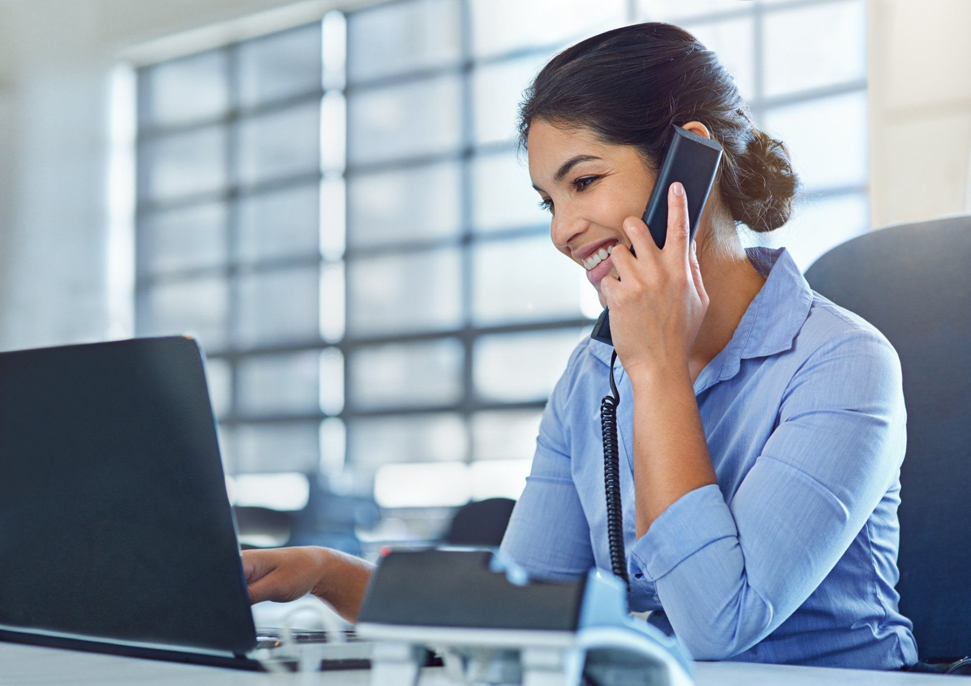 A woman is sitting at a desk talking on a cell phone while using a laptop computer.