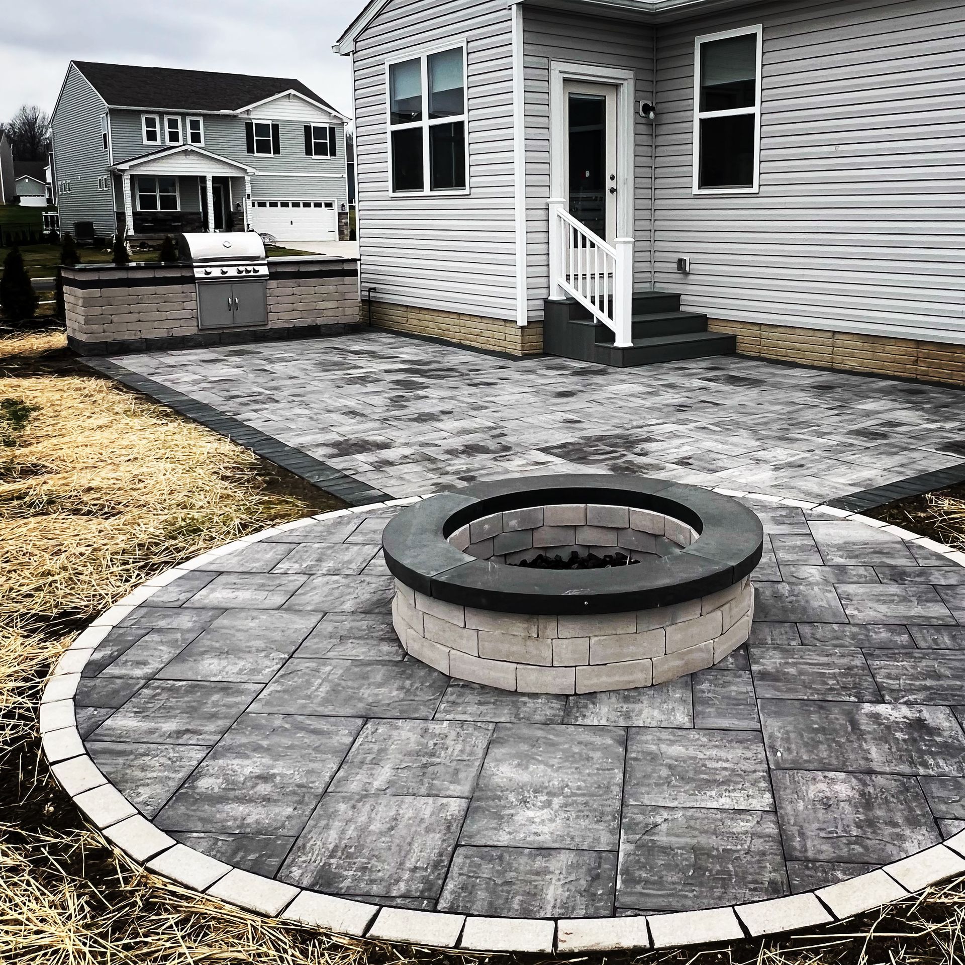 Backyard patio with fire pit, grill, and steps leading to a house with gray siding.