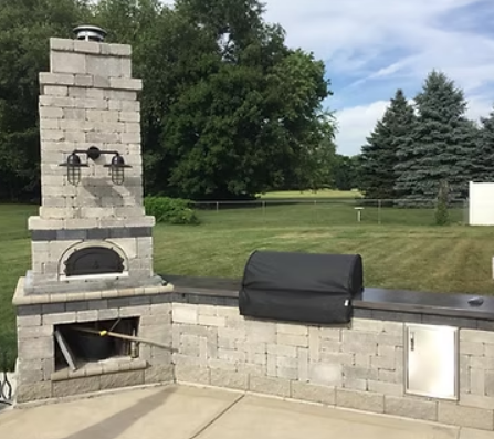 Stone outdoor pizza oven and grill in a backyard with trees.