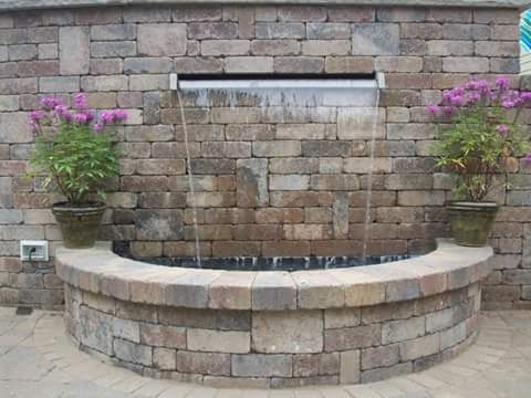 Stone wall water fountain with cascading water and potted flowers.