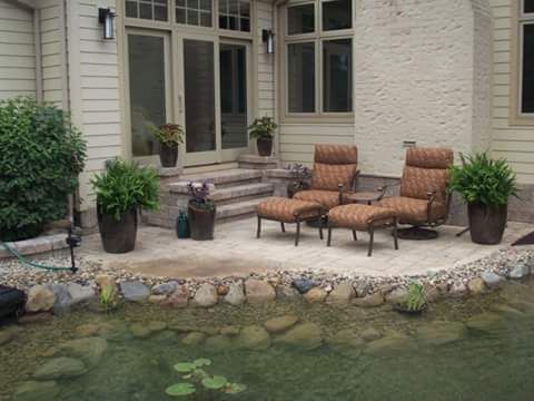 Patio with seating next to a pond. Ferns in pots, beige stone patio, light-colored siding, and a two-chair set.