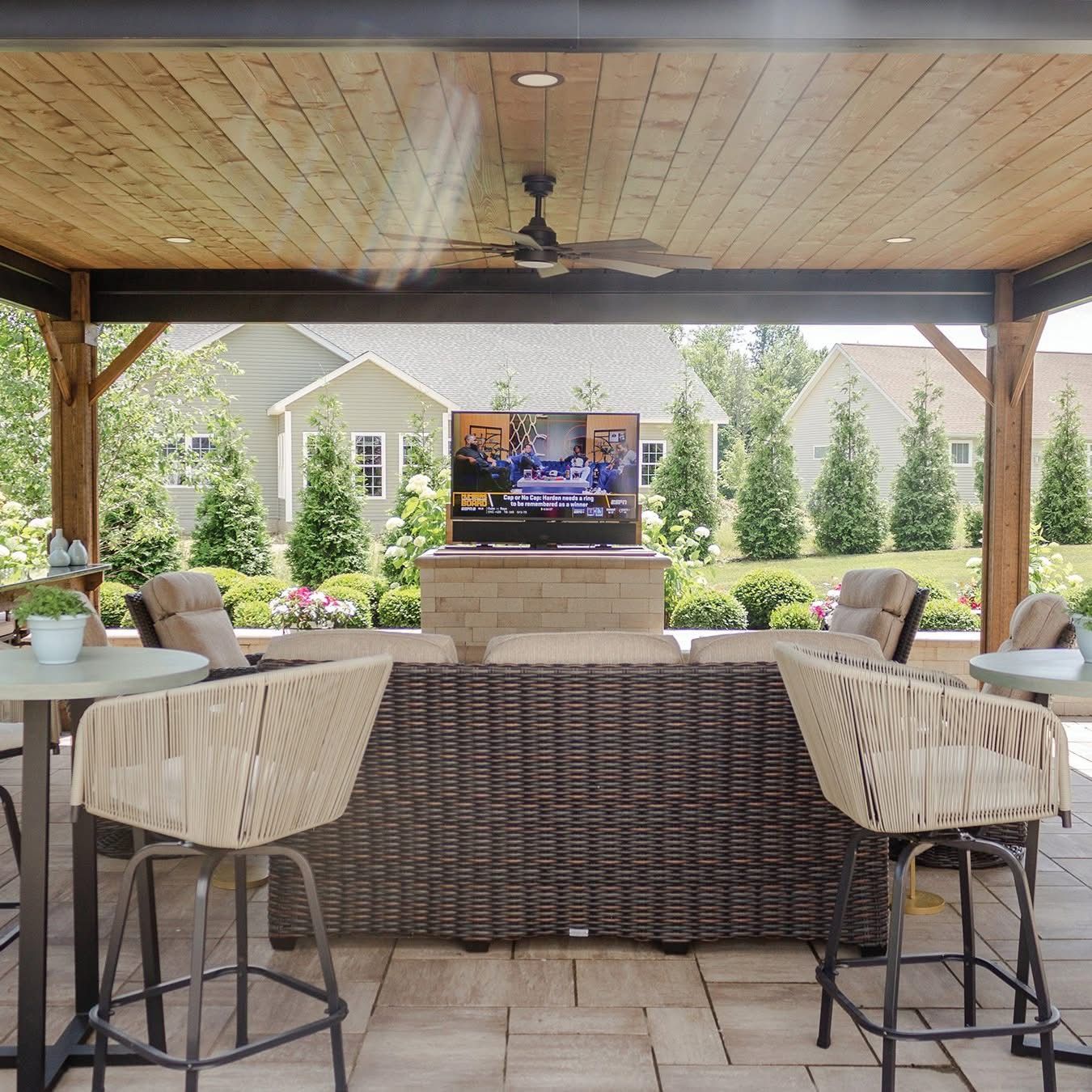 Outdoor patio with seating, TV, and wood ceiling, overlooking a suburban backyard.