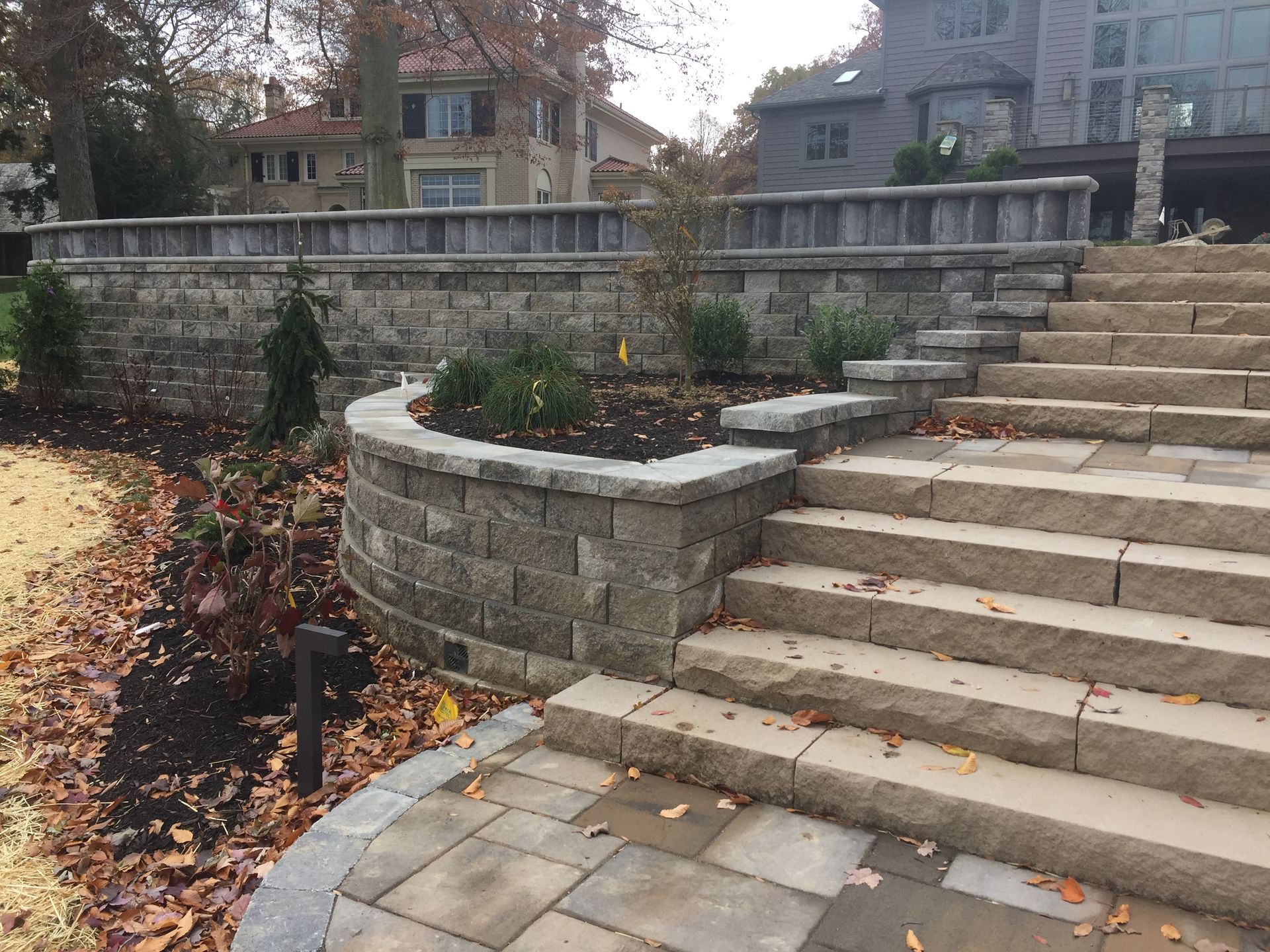 Stone steps and retaining walls in a landscaped yard. Brown bricks, fall foliage, and a house in the background.