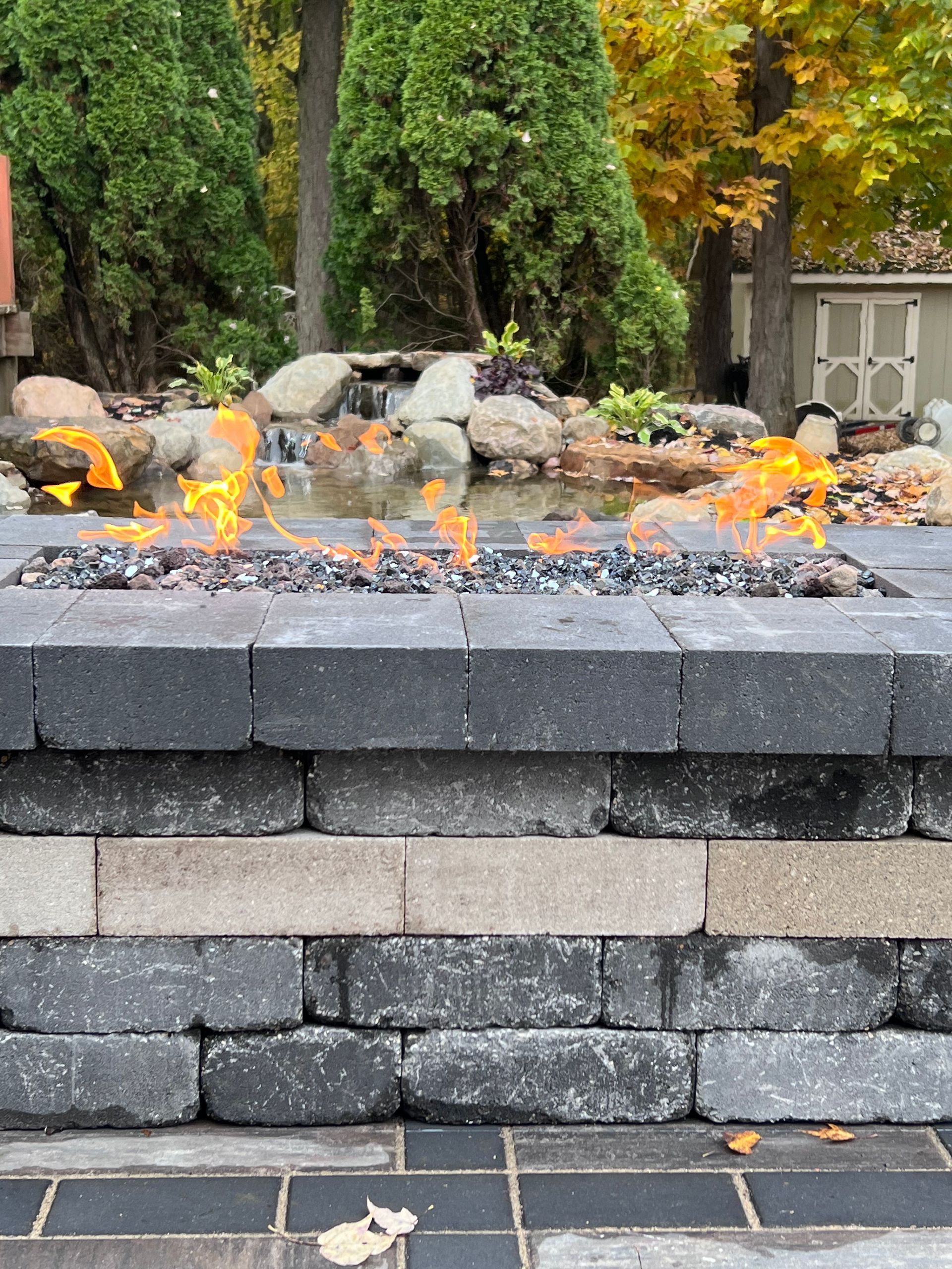 Fire pit with flames against a background of a water feature and trees.
