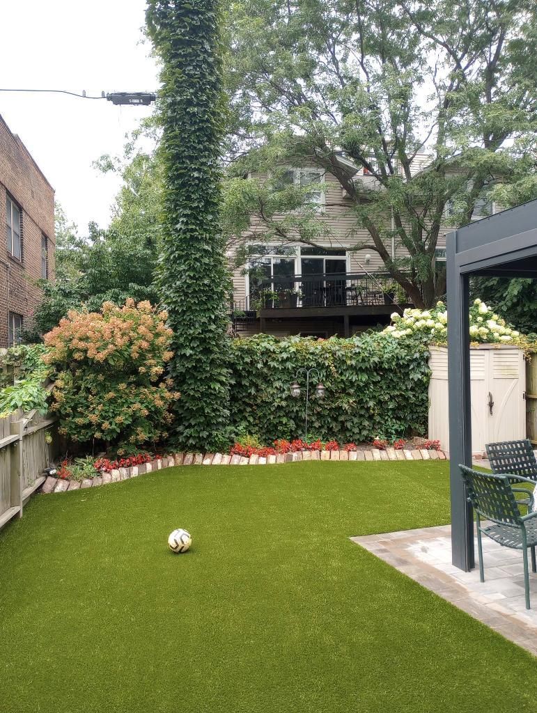 A green backyard with a ball on turf, surrounded by plants, a brick building, and a pergola.