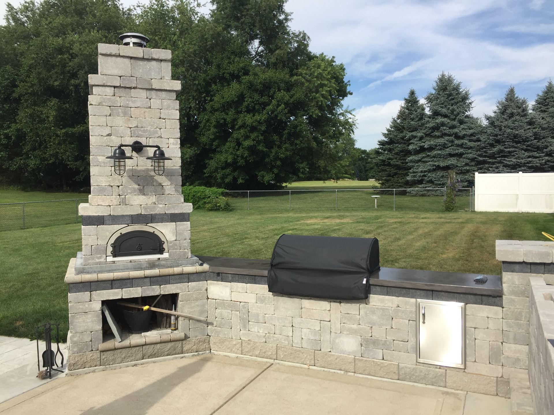 Outdoor kitchen with stone pizza oven, grill, and stainless steel cabinet on a patio, green lawn in background.
