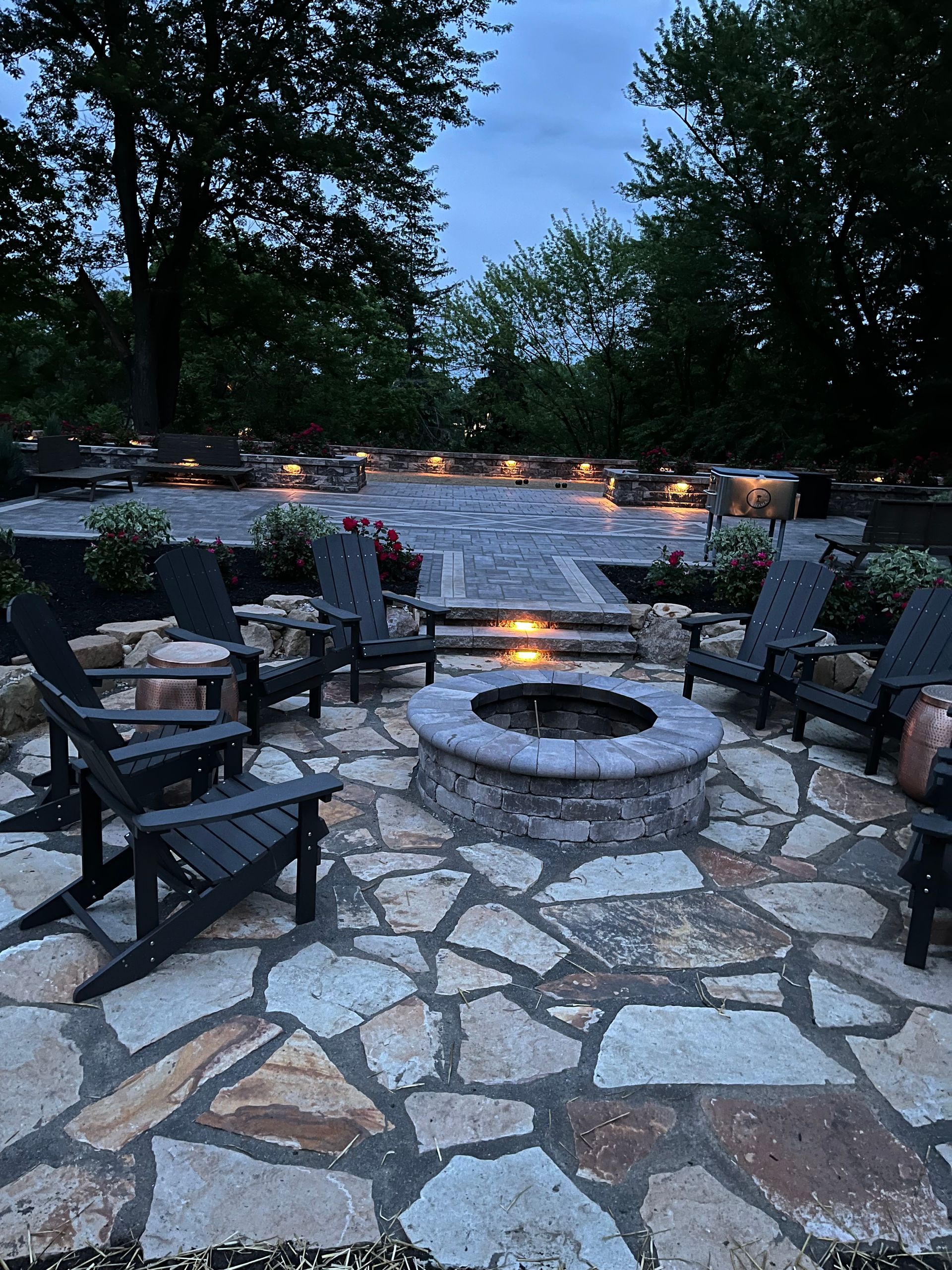 Stone patio with fire pit, Adirondack chairs, and landscaping, lit at dusk.