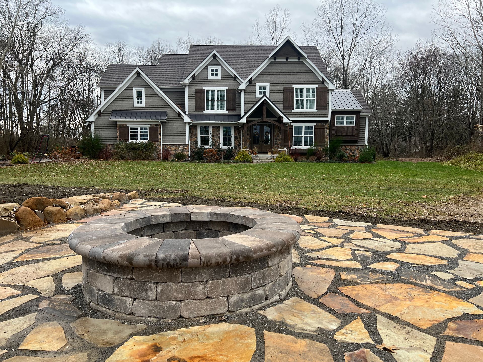 Fire pit on stone patio with a large house in the background, surrounded by grass and trees.