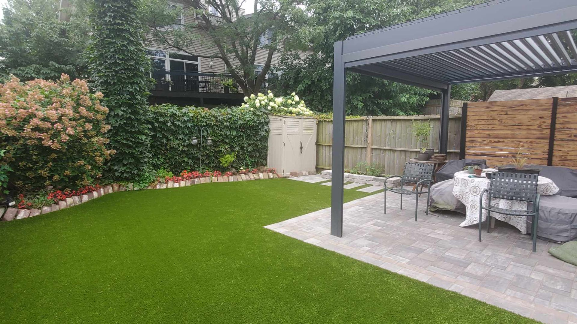 Backyard with green lawn, patio, seating, and pergola, bordered by plants and a fence.