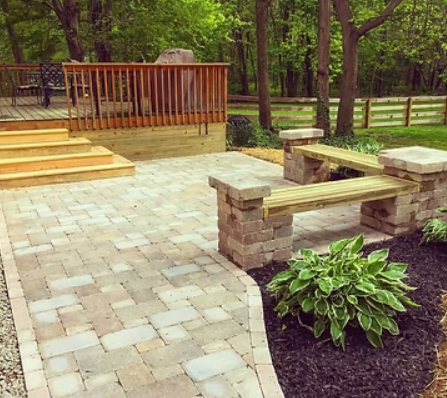 Brick patio with wood benches, deck, steps, and green landscaping.