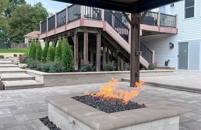 Fire pit on stone patio, with a raised deck and landscaping in the background.
