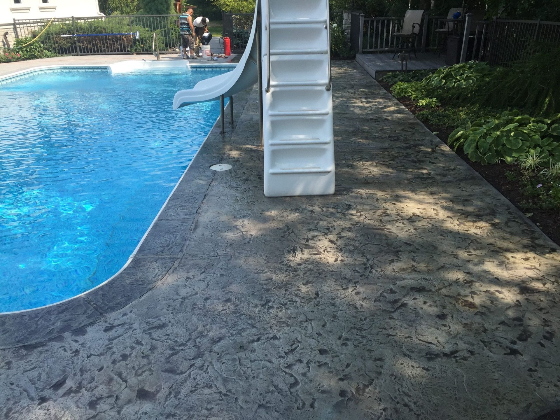 Swimming pool deck with a white slide and blue water beside a shaded paved path