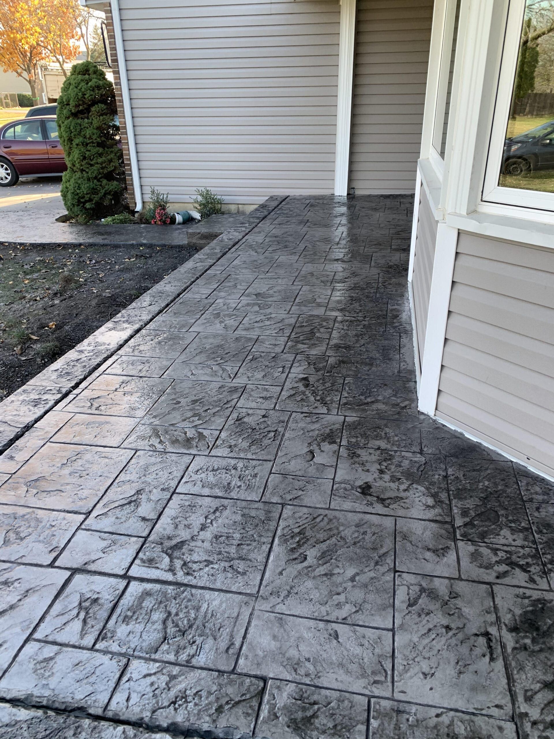 Stamped concrete walkway beside a house with a door and window, leading toward a driveway and shrubs.