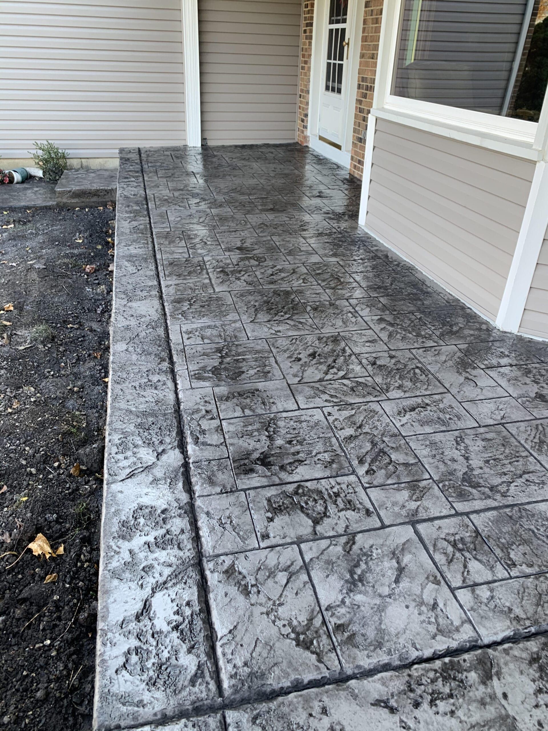 Stamped concrete patio walkway beside a house with dark stain patterns and a door at the end