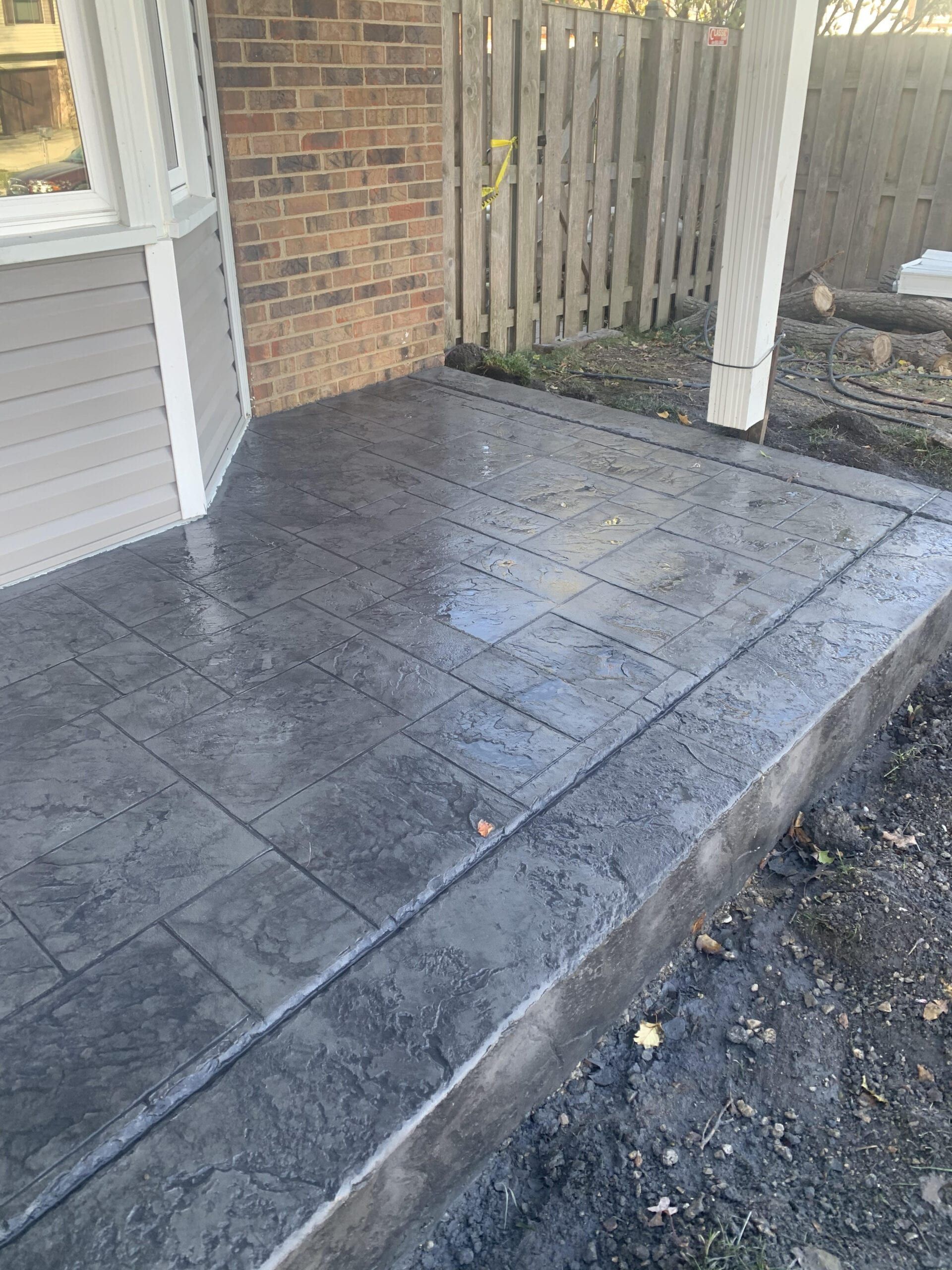 Dark gray tiled patio beside a brick house and wooden fence, with a gravel edge.