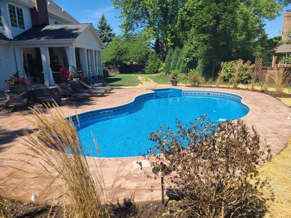 Backyard swimming pool beside a house, surrounded by a stone patio and trees on a sunny day