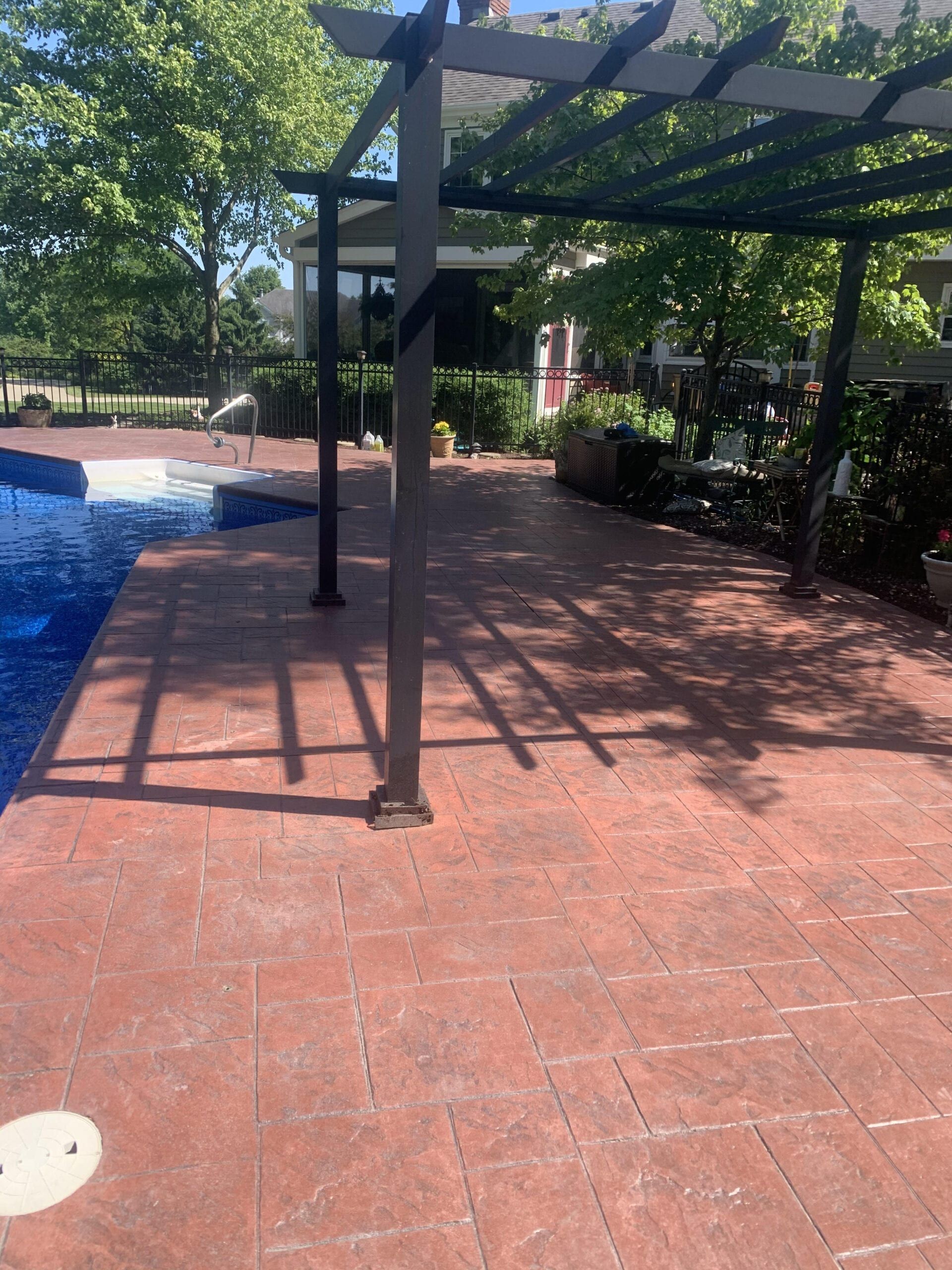 Shaded poolside patio with red brick pavers, black pergola, and blue water on the left
