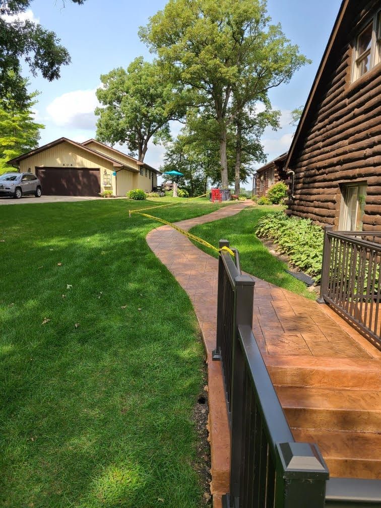 Wooded hillside walkway with a cabin, green lawn, and log house under a sunny sky
