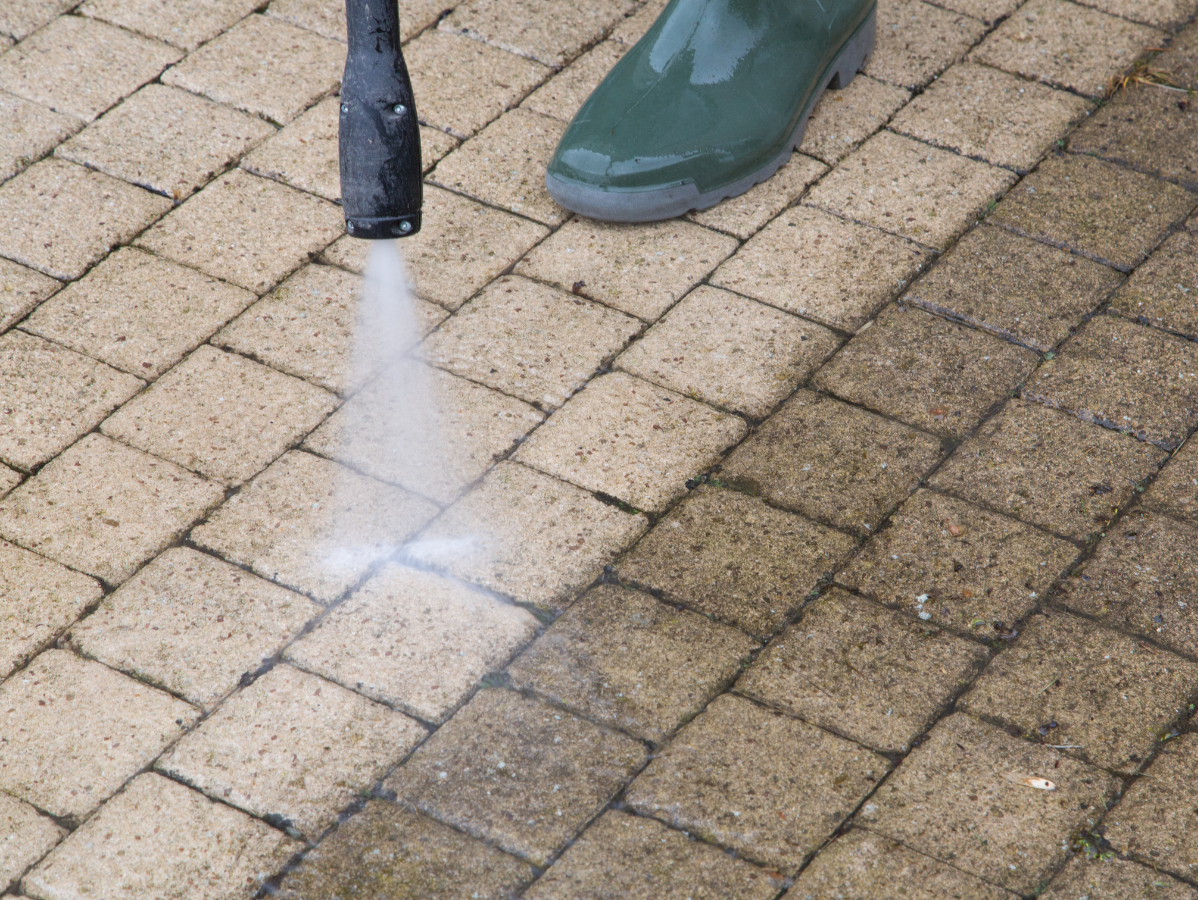 Person pressure-washing a brick patio with a black nozzle and white spray.
