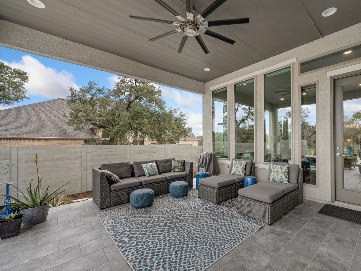 Covered patio with gray outdoor seating, blue rug, and ceiling fan beside large glass doors.