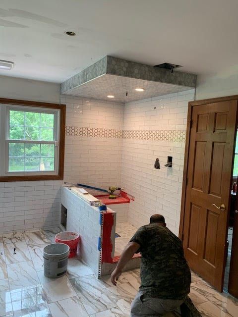 Worker installing floor tile in a tiled bathroom with a vanity, window, and wooden door.